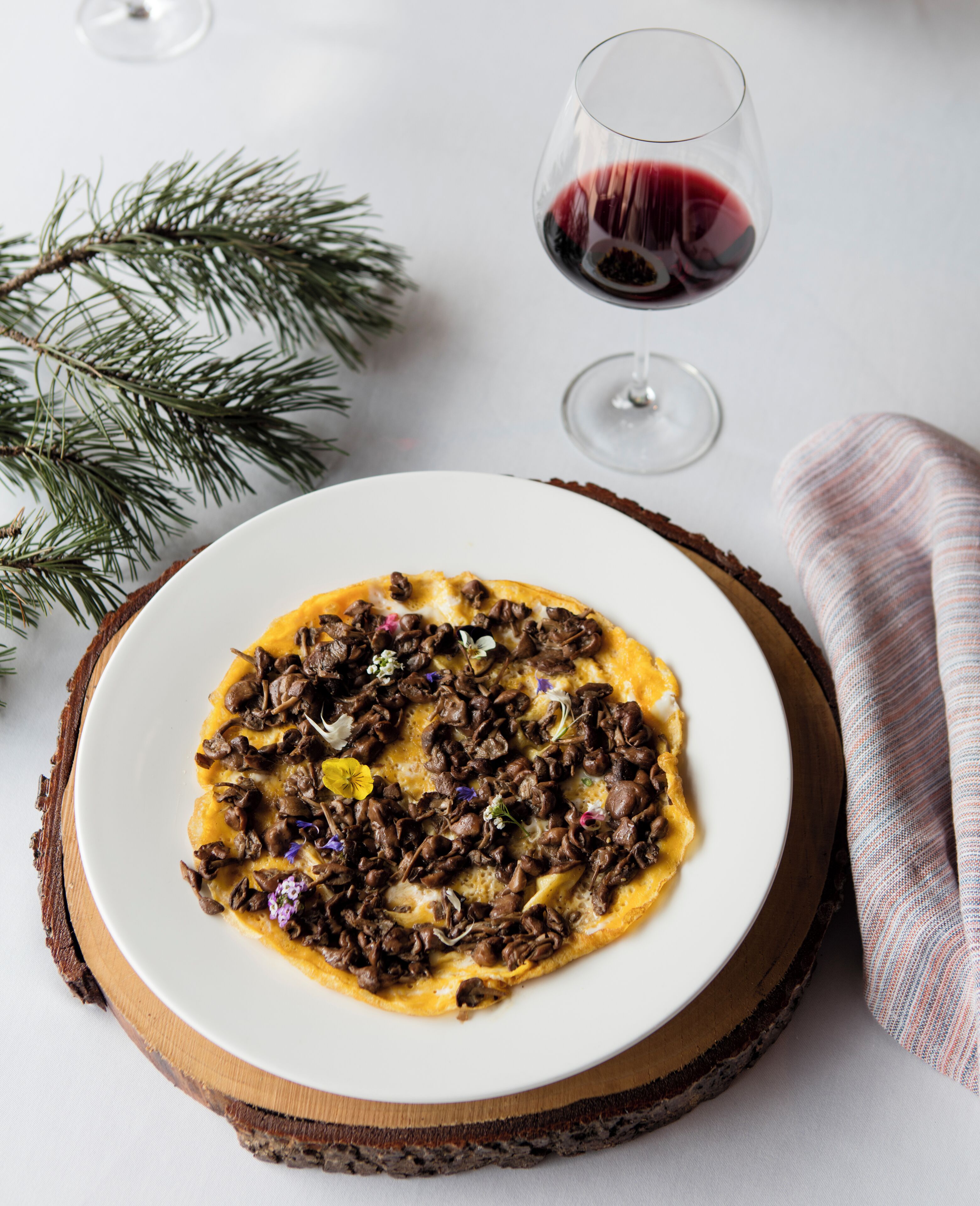 A plate of food on a wooden board with a glass of red wine and a pine branch in the background.