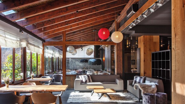 Un interior moderno con vigas de madera, un sofá, una zona de comedor y ventanas grandes.