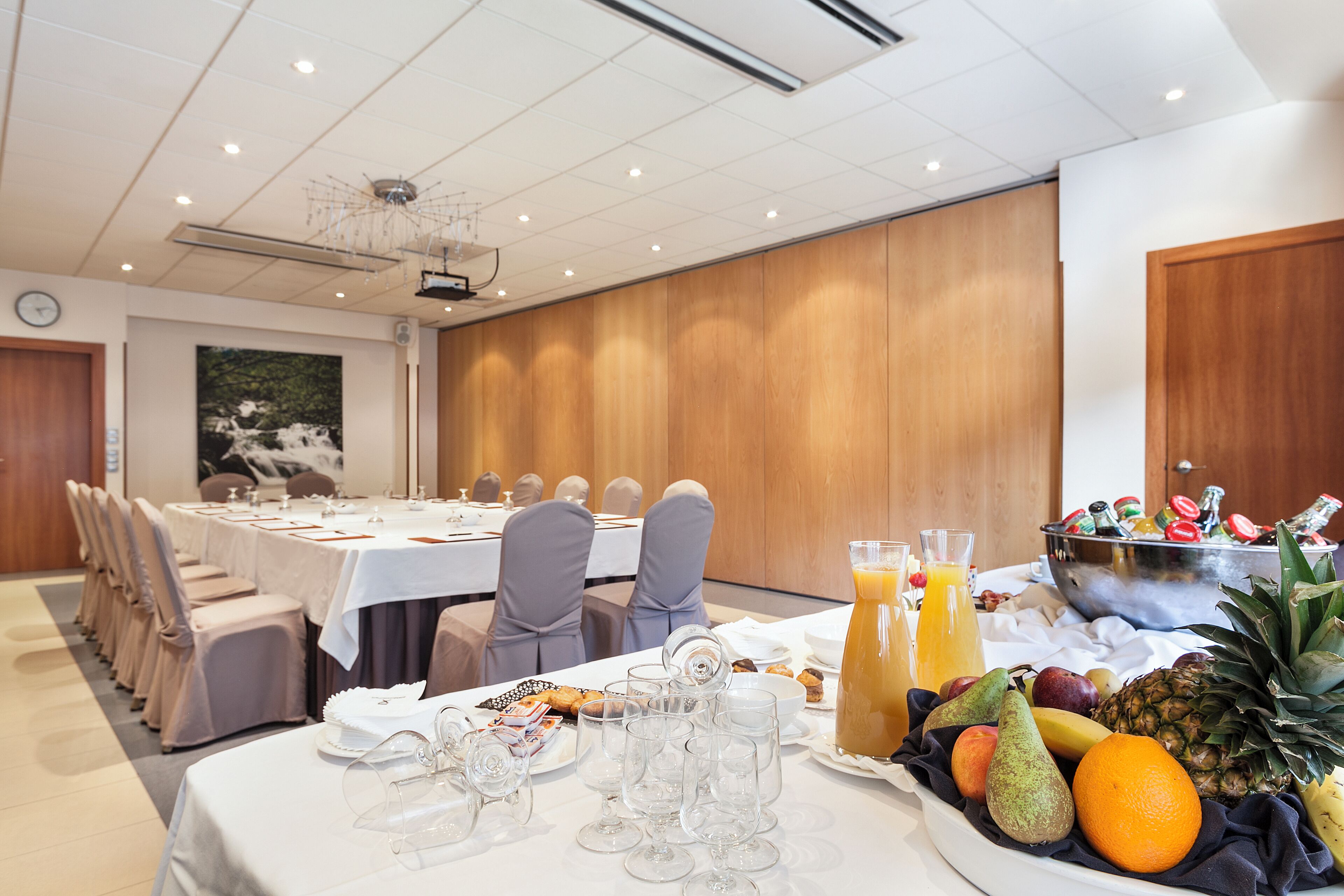 Un salón de conferencias con una mesa larga preparada para una reunión, una presentación de frutas y jugos, y una mesa de buffet con diversos bocadillos.