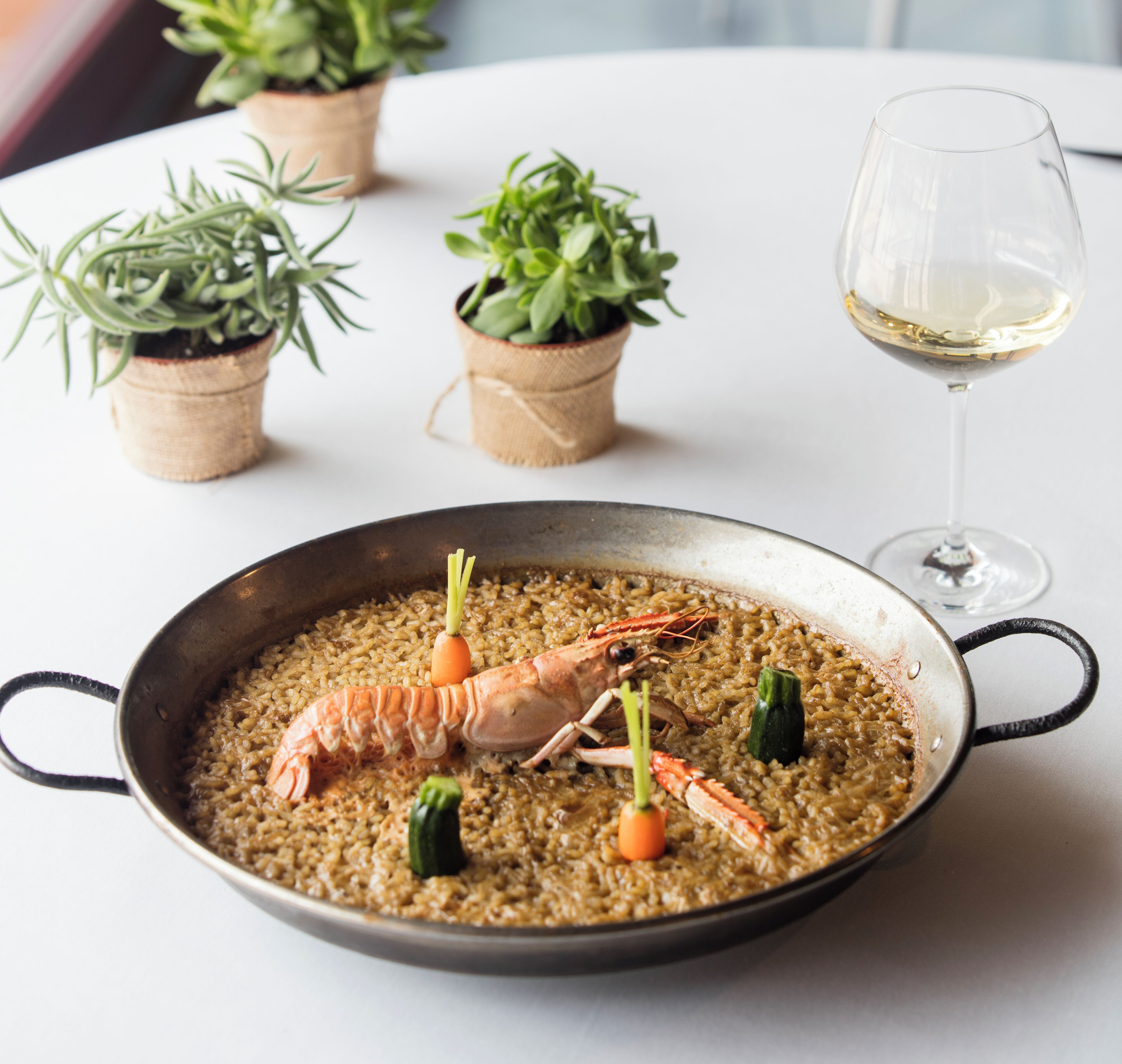 A seafood dish in a pan, a glass of white wine, and small potted plants on a table.