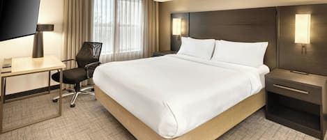 A hotel room with a large bed, a desk with a flat-screen TV, a chair, and a nightstand with a lamp.
