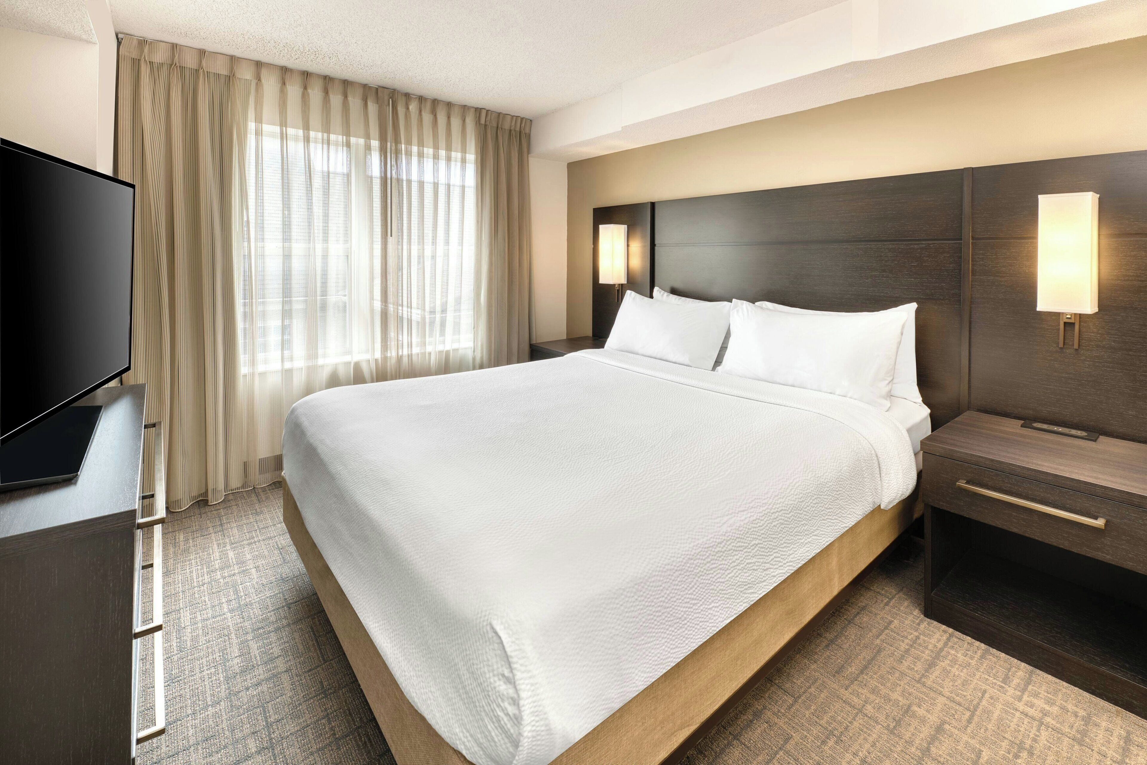A hotel room with a large bed, a flat-screen TV, a nightstand, a lamp, and a window with curtains.