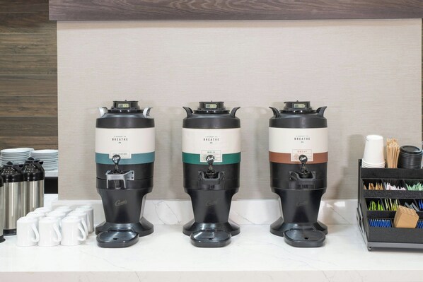 Tres cafeteras con la etiqueta 'BREATHE' sobre una encimera, junto a tazas de café y un recipiente de almacenamiento.