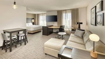A hotel room with a bed, a desk, a sofa, a dining table with chairs, a TV, and framed pictures on the wall.