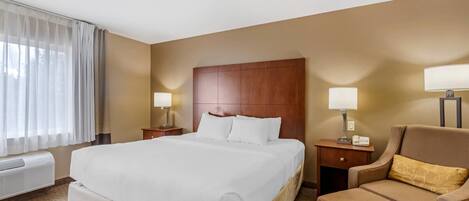 A hotel room with a large bed, a wooden headboard, two bedside tables with lamps, a brown armchair, and a window with sheer curtains.