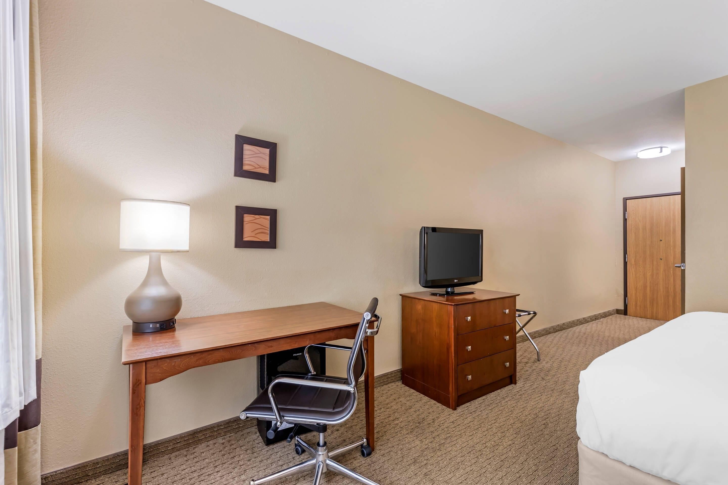 A hotel room with a wooden desk, a chair, a television, a dresser, and a bed.