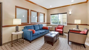 A hotel room with a blue sofa, a red chair, a brown ottoman, and a patterned carpet.