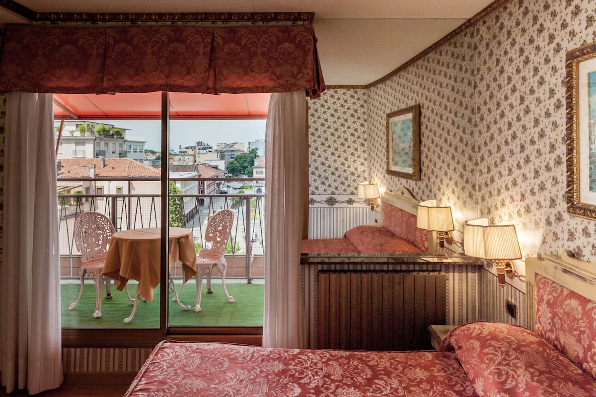 Habitación de hotel con balcón, cama con ropa de cama estampada, una mesita con sillas y una mesita de noche con lámparas.