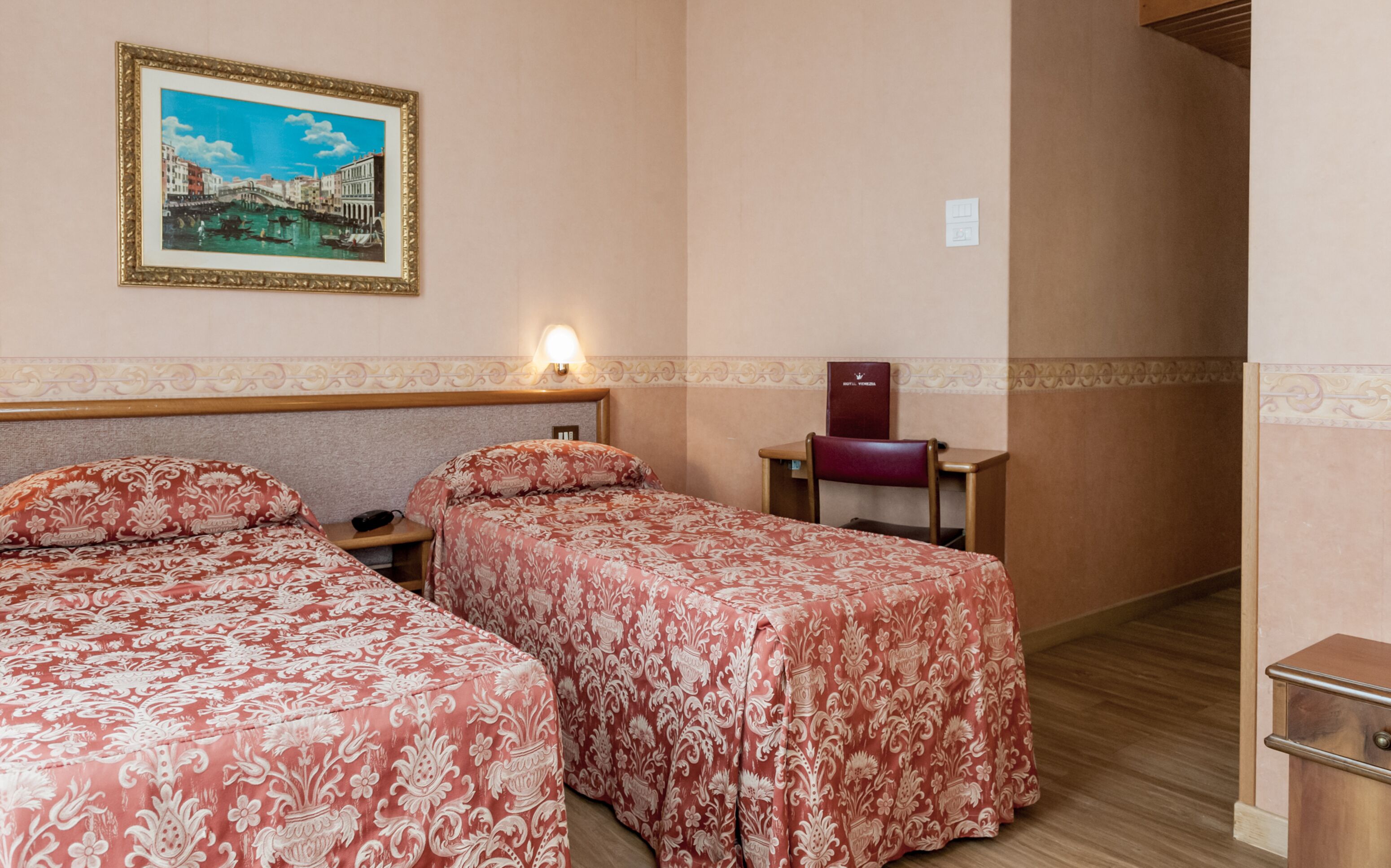 Habitación de hotel con dos camas, un escritorio, una silla, una mesita de noche, una lámpara y un cuadro en la pared.