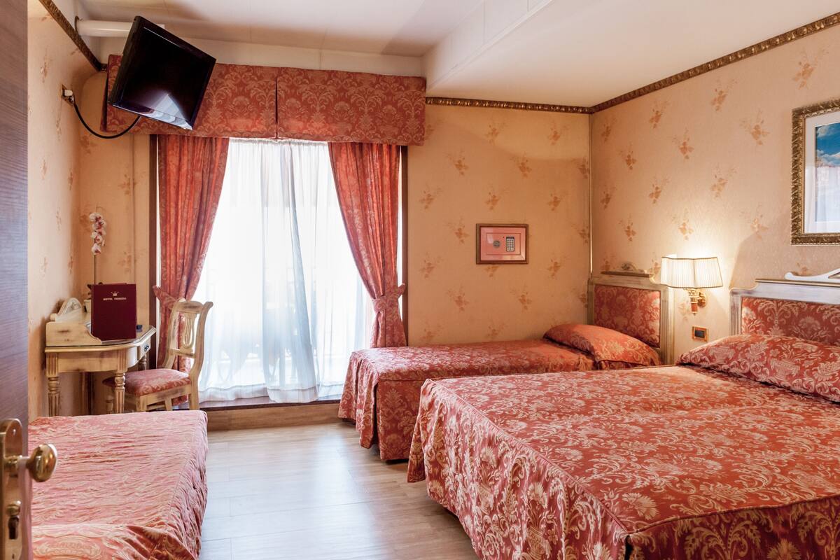 Habitación de hotel con tres camas, un escritorio y una silla, un televisor en la pared y una ventana con cortinas.