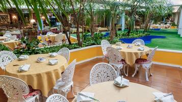 An indoor dining area with round tables and white chairs, surrounded by greenery and a small pool.
