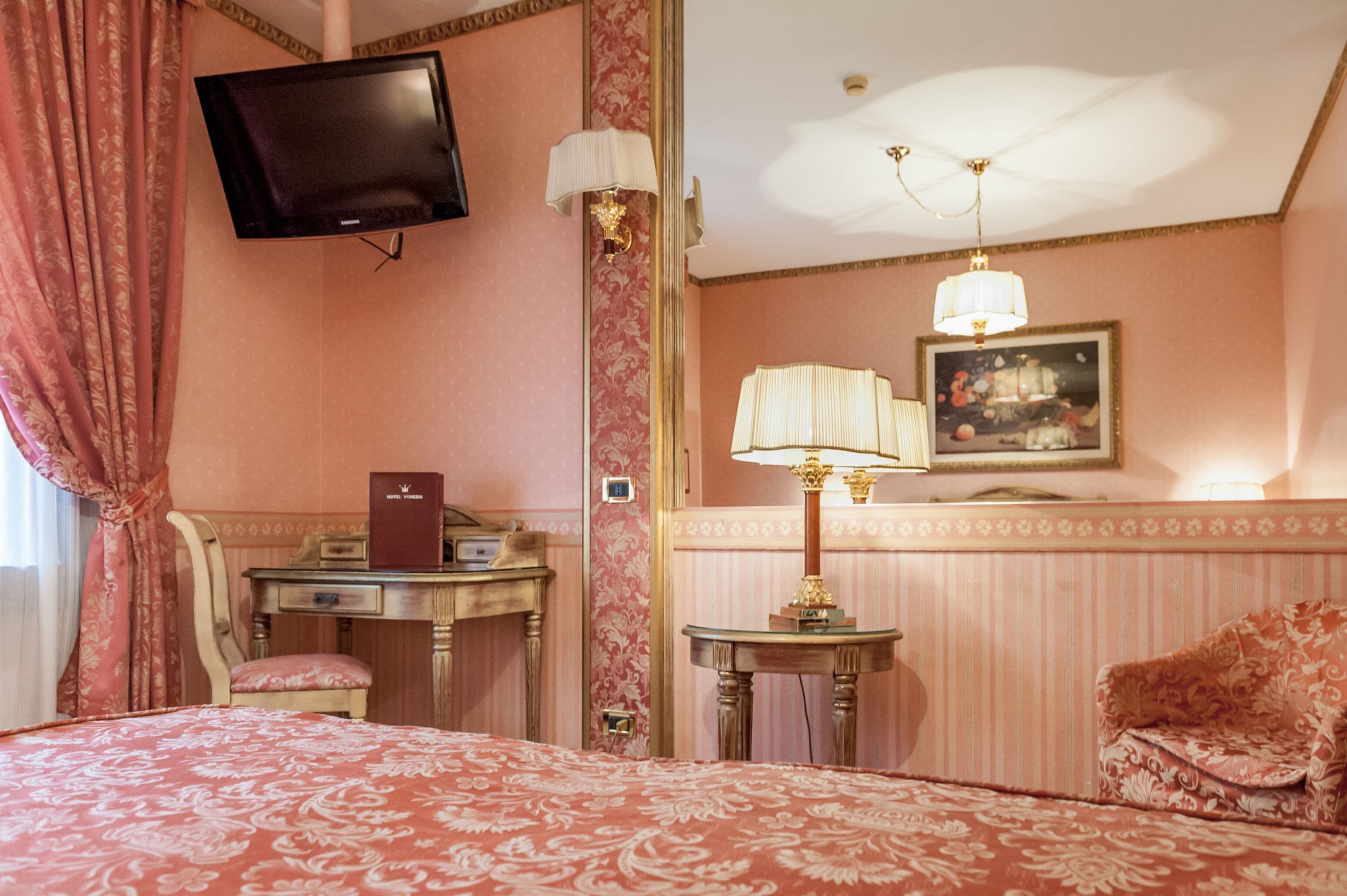 Habitación de hotel con cama, escritorio, silla, televisión, lámpara y un cuadro en la pared.