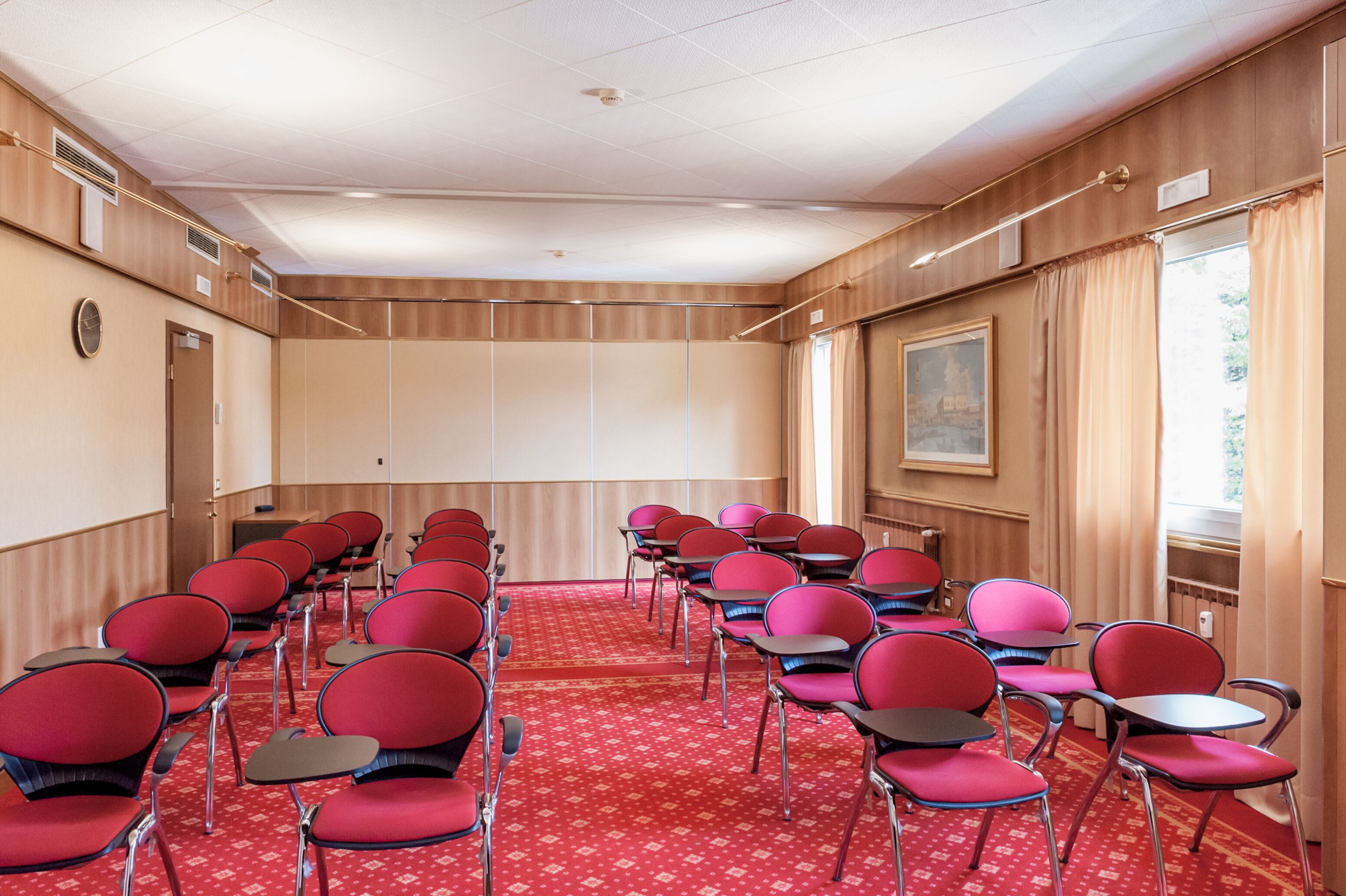 A conference room with rows of red chairs and tables, a carpeted floor, wooden panel walls, and a large window with curtains.