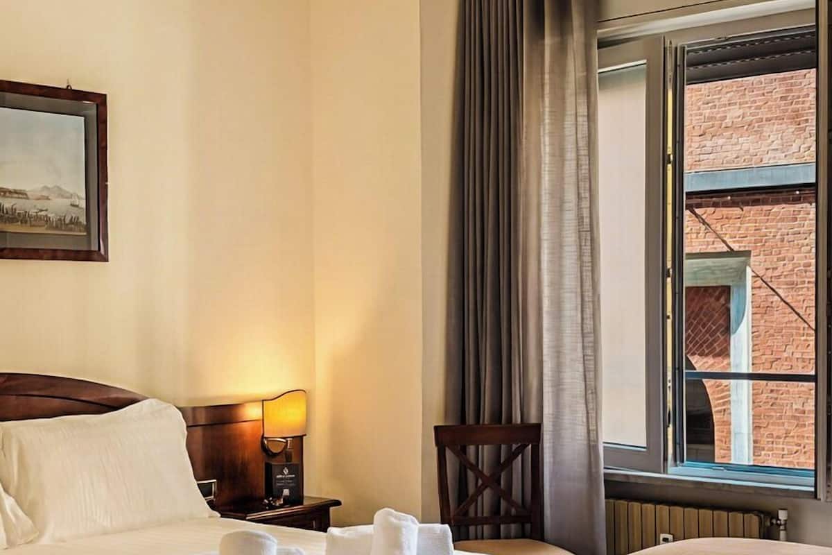 A hotel room with two beds, a nightstand with a lamp, a chair, and a window with curtains.