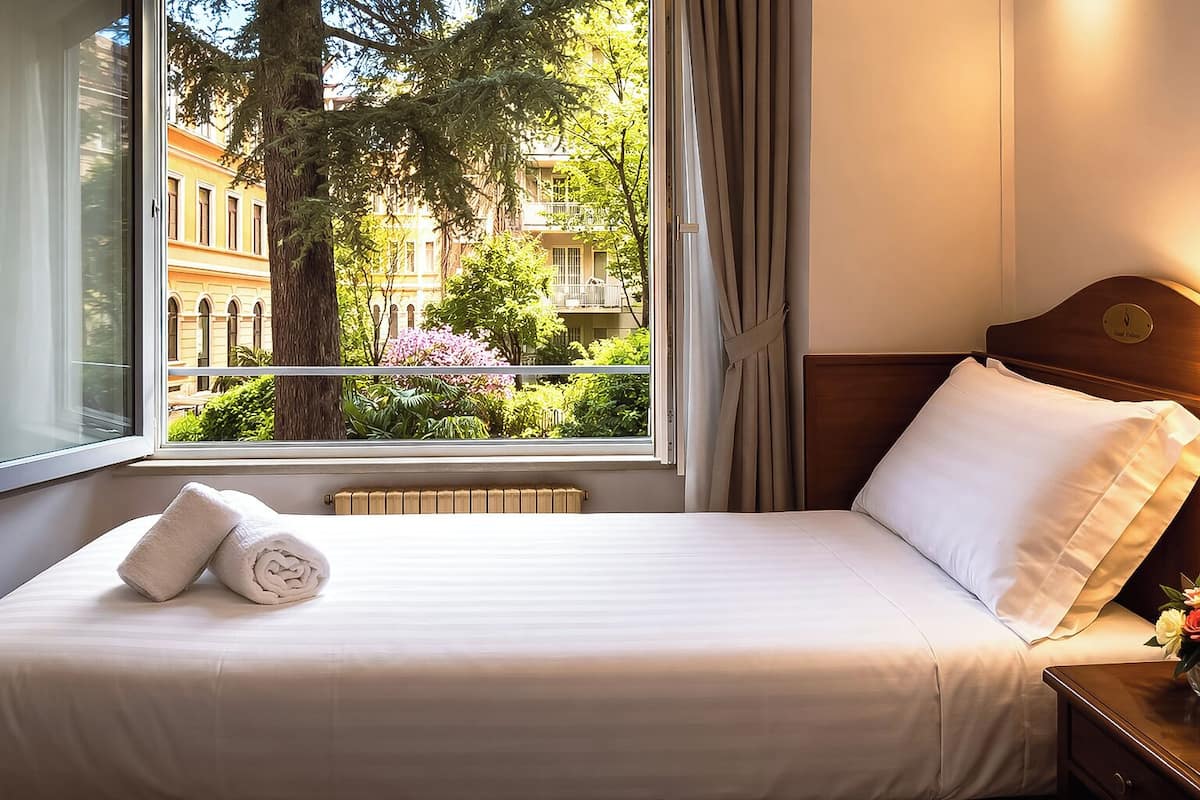 A hotel room with a bed, a window with curtains, a radiator, and a view of buildings and trees.
