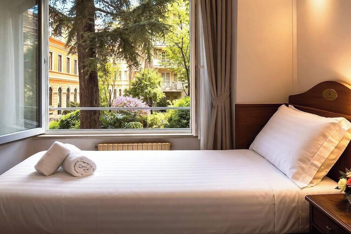 A hotel room with a bed, a window with a view of buildings and trees, a radiator, and a nightstand with a lamp.