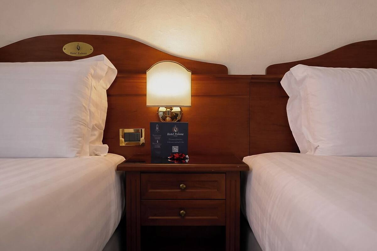 A hotel room with two beds, a nightstand with a lamp, a clock, and a framed welcome sign.