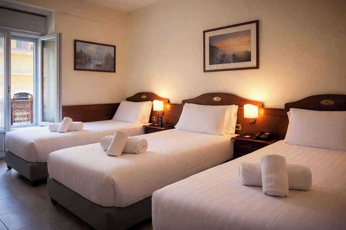 A hotel room with three beds, each with a pillow and a rolled towel. There are two wall-mounted lamps on each nightstand.