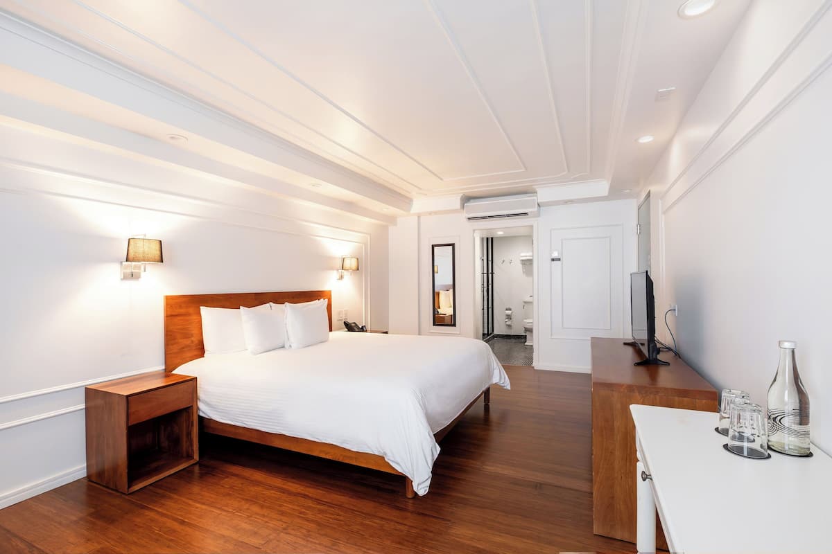 A modern hotel room with a large bed, wooden nightstands, a desk with a bottle and glasses, and a TV.