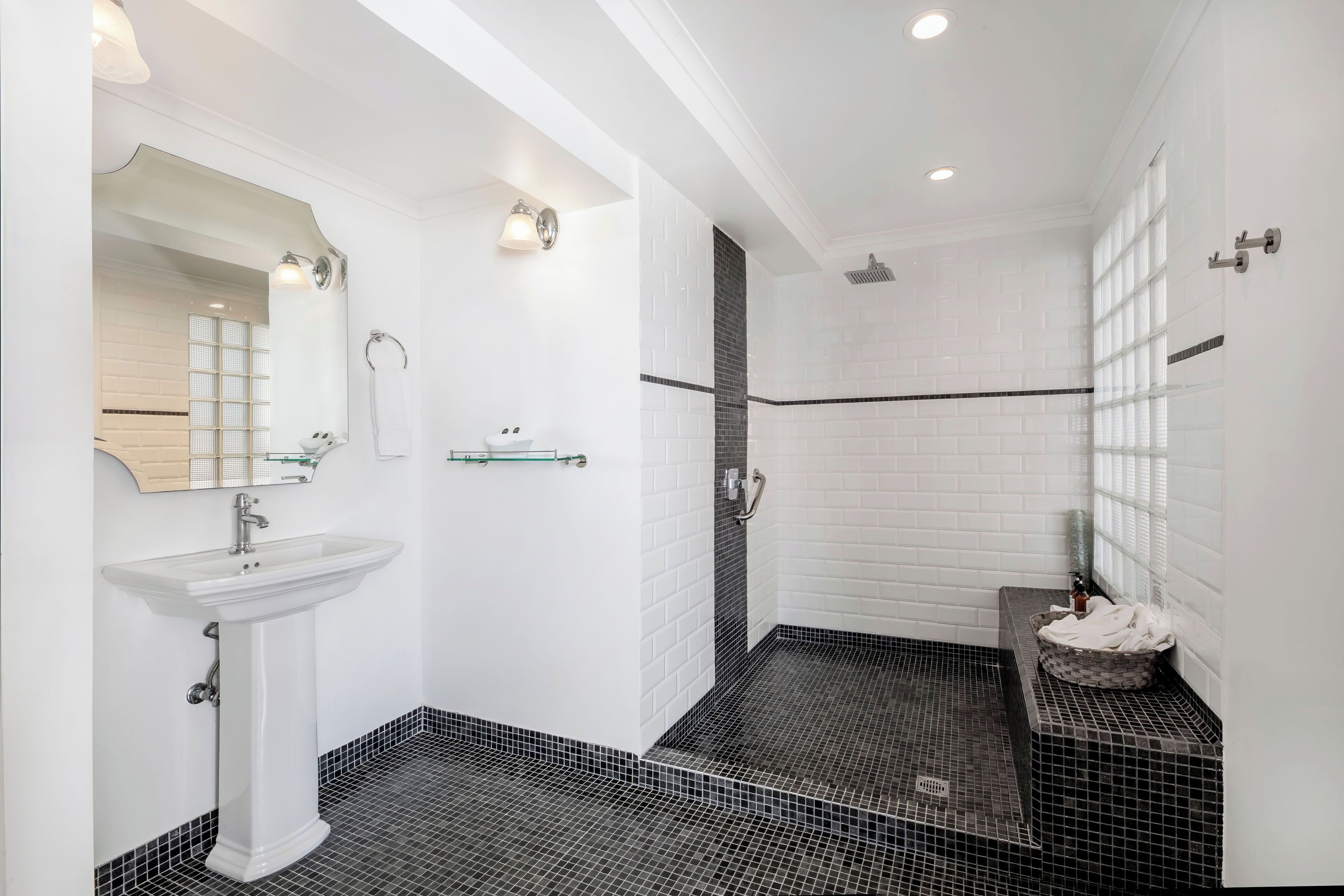 A modern bathroom with a white pedestal sink, a glass shelf, a black and white tiled floor, and a shower area.