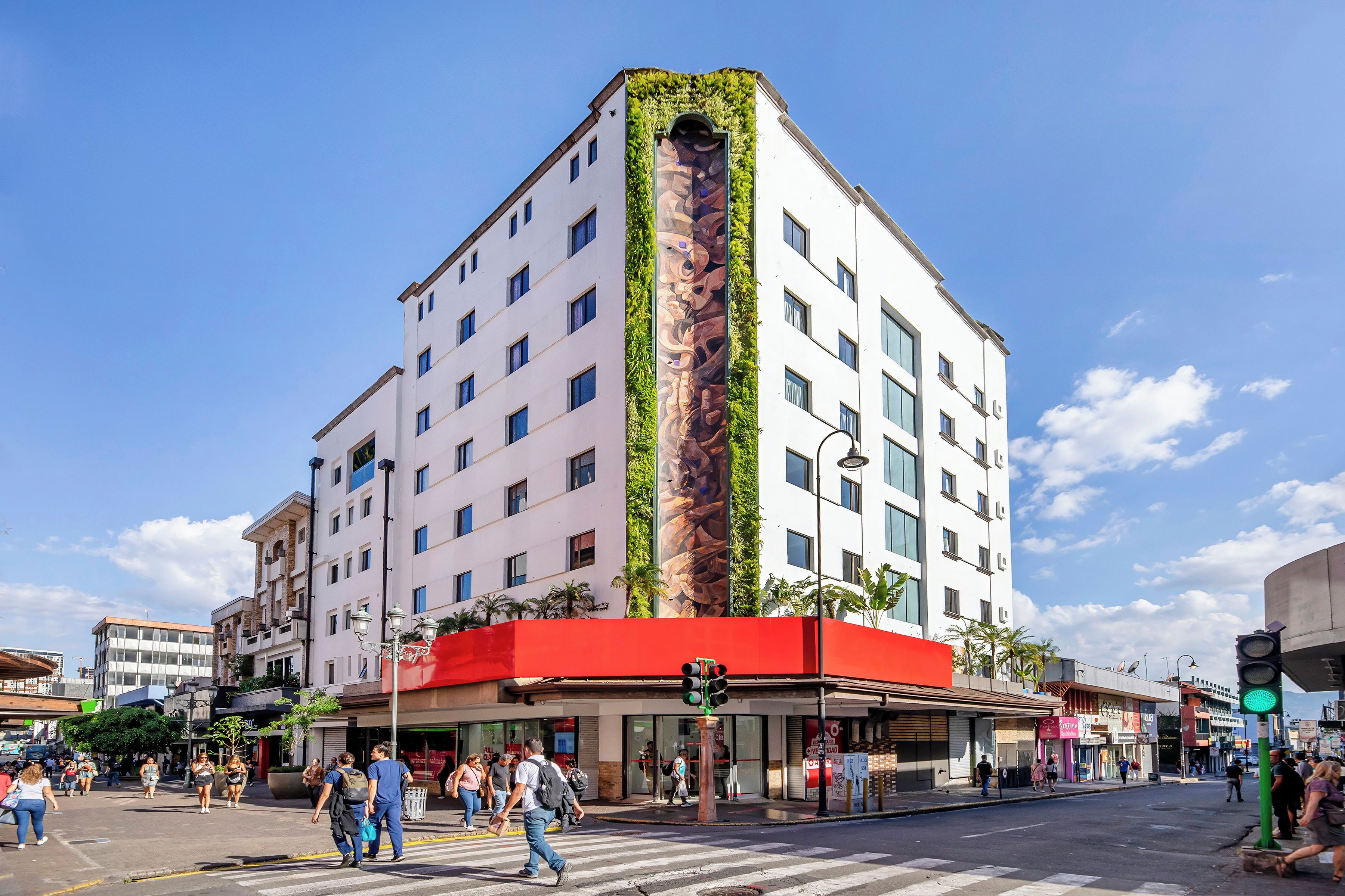 A multi-story hotel with a red awning, a vertical garden on the facade, and a pedestrian crossing.