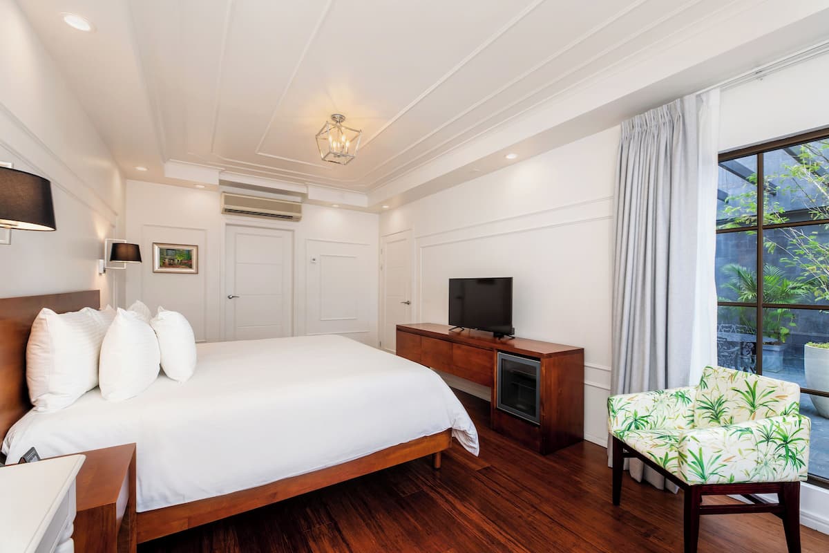 A bedroom with a wooden bed, a TV on a wooden cabinet, a chair with a leaf pattern, and a window with a view of greenery.