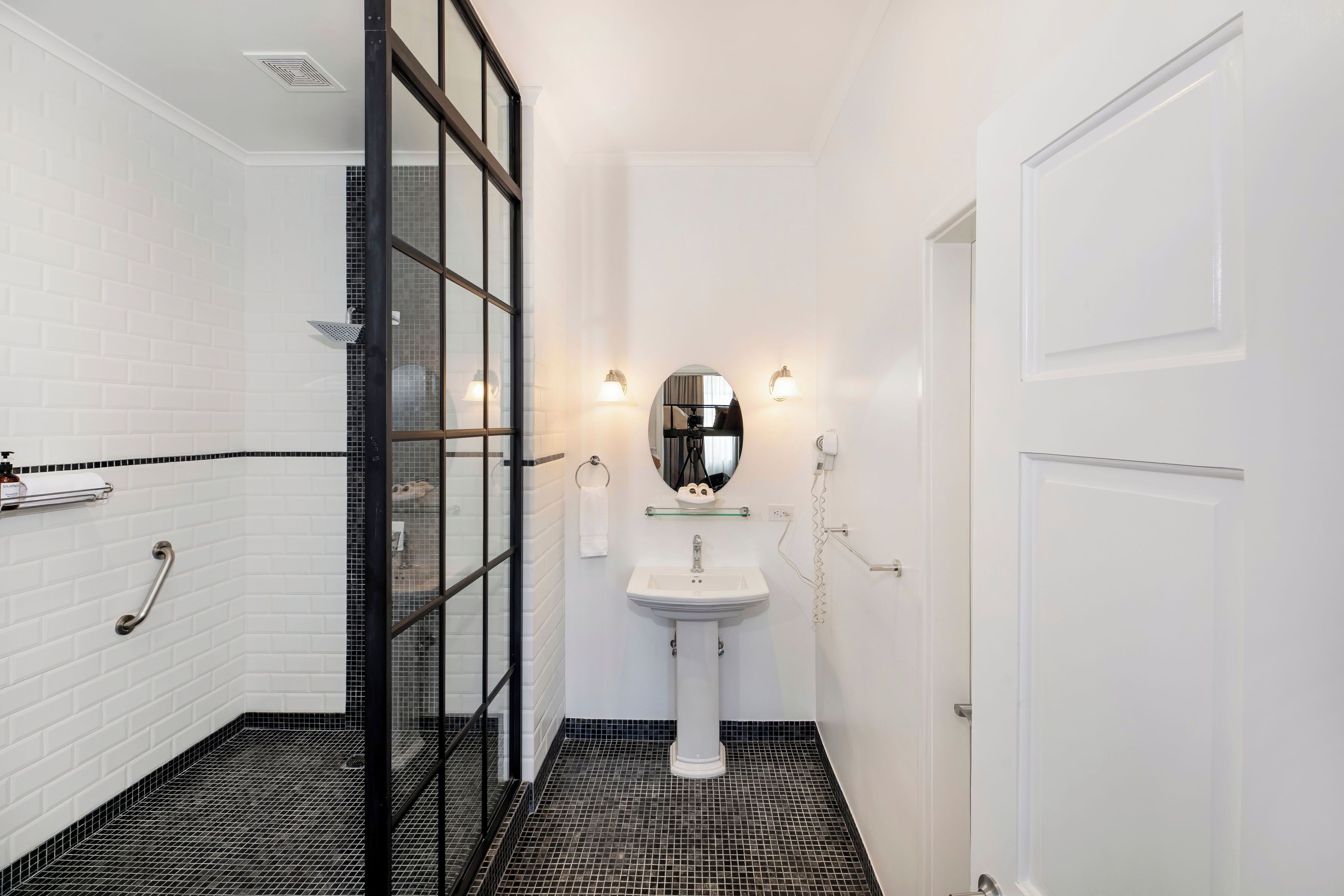 A bathroom with white subway tiles, a black and white tiled floor, a glass shower enclosure, a white pedestal sink, and a mirror above the sink.