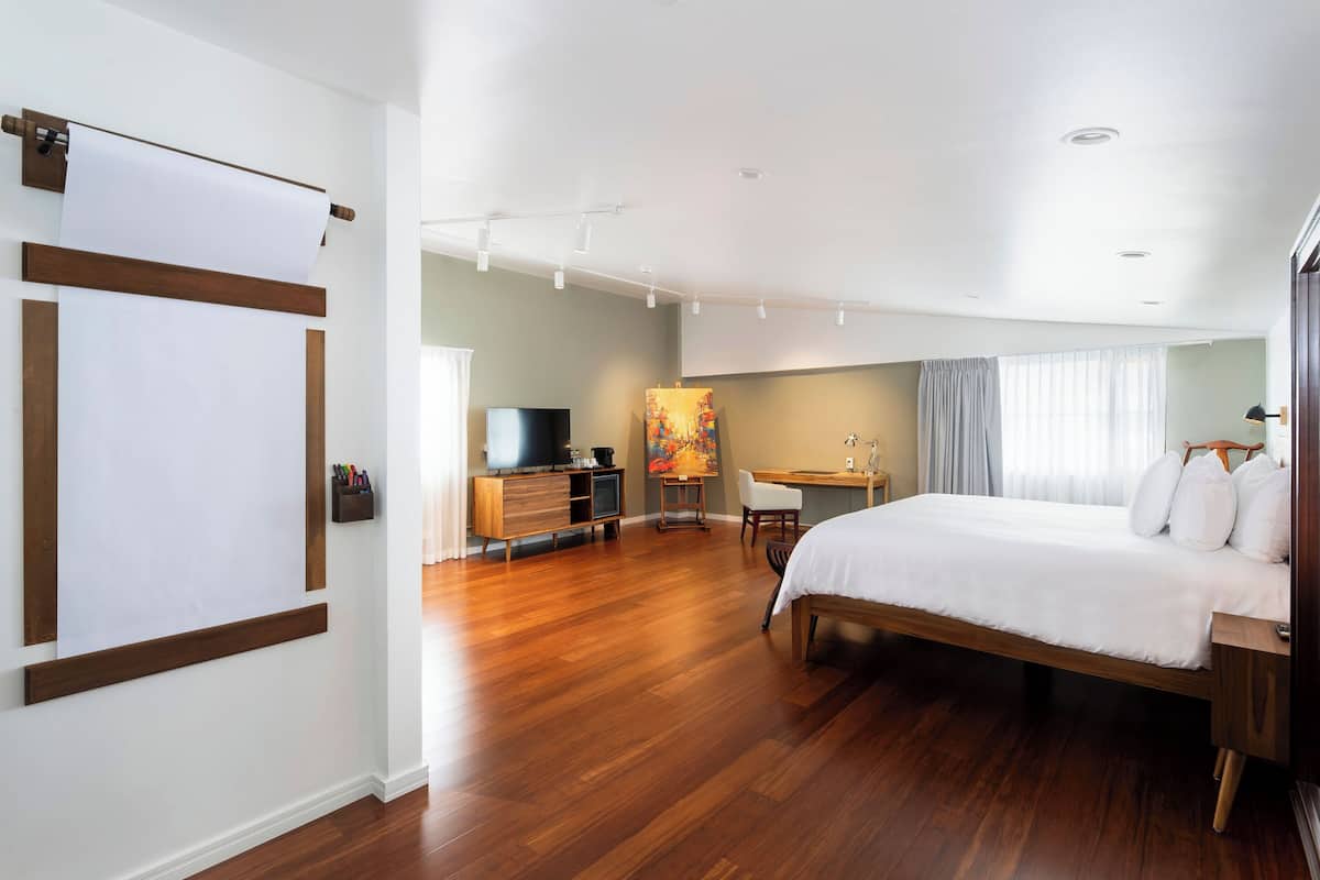 A modern hotel room with a wooden floor, a bed with white bedding, a desk with a chair, a television, and a painting on the wall.