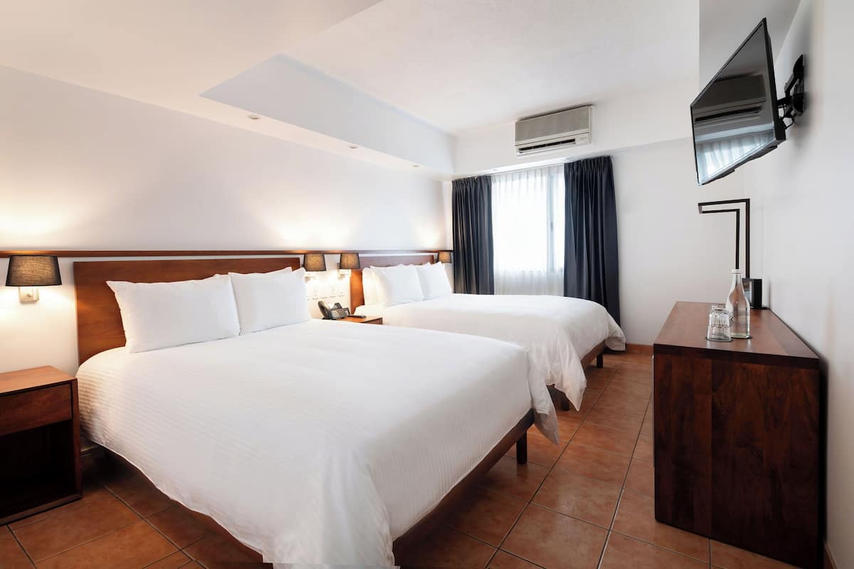 A hotel room with two beds, a wooden headboard, a flat-screen TV mounted on the wall, a bedside table with a lamp, and a small wooden cabinet.