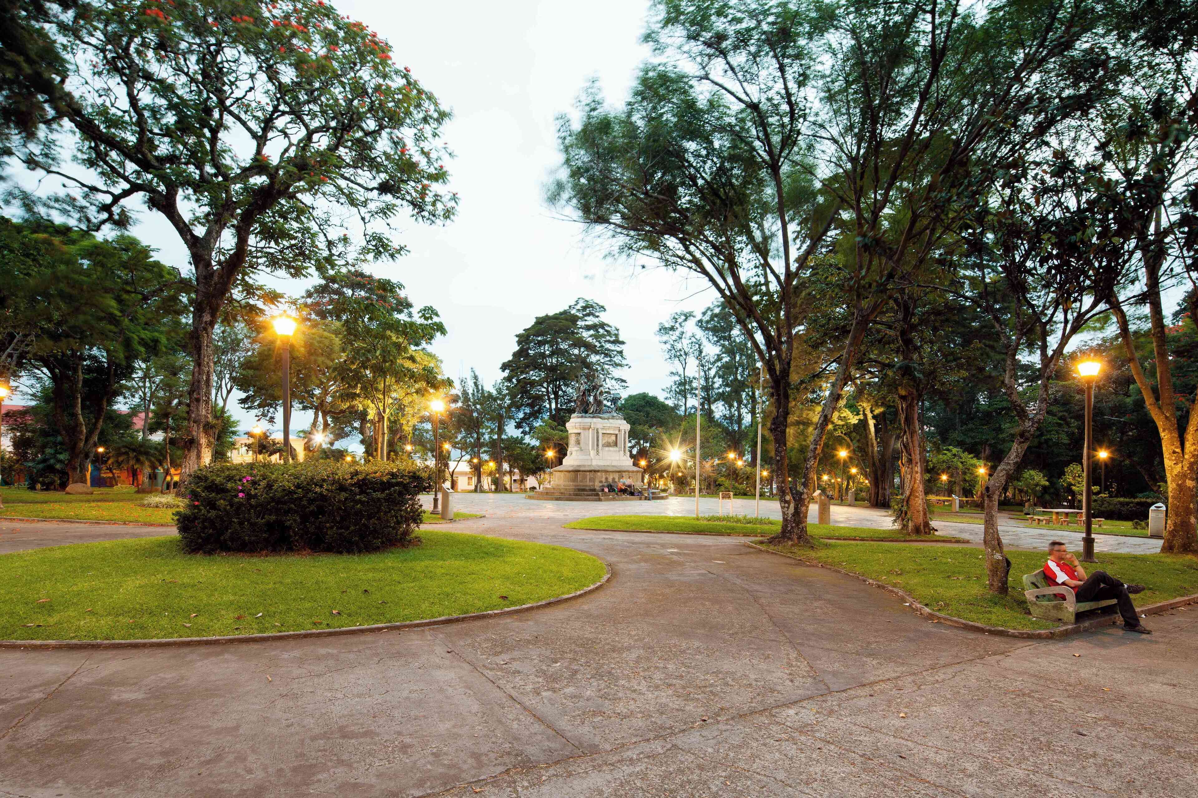 Um parque com um monumento central, caminhos, bancos e árvores.