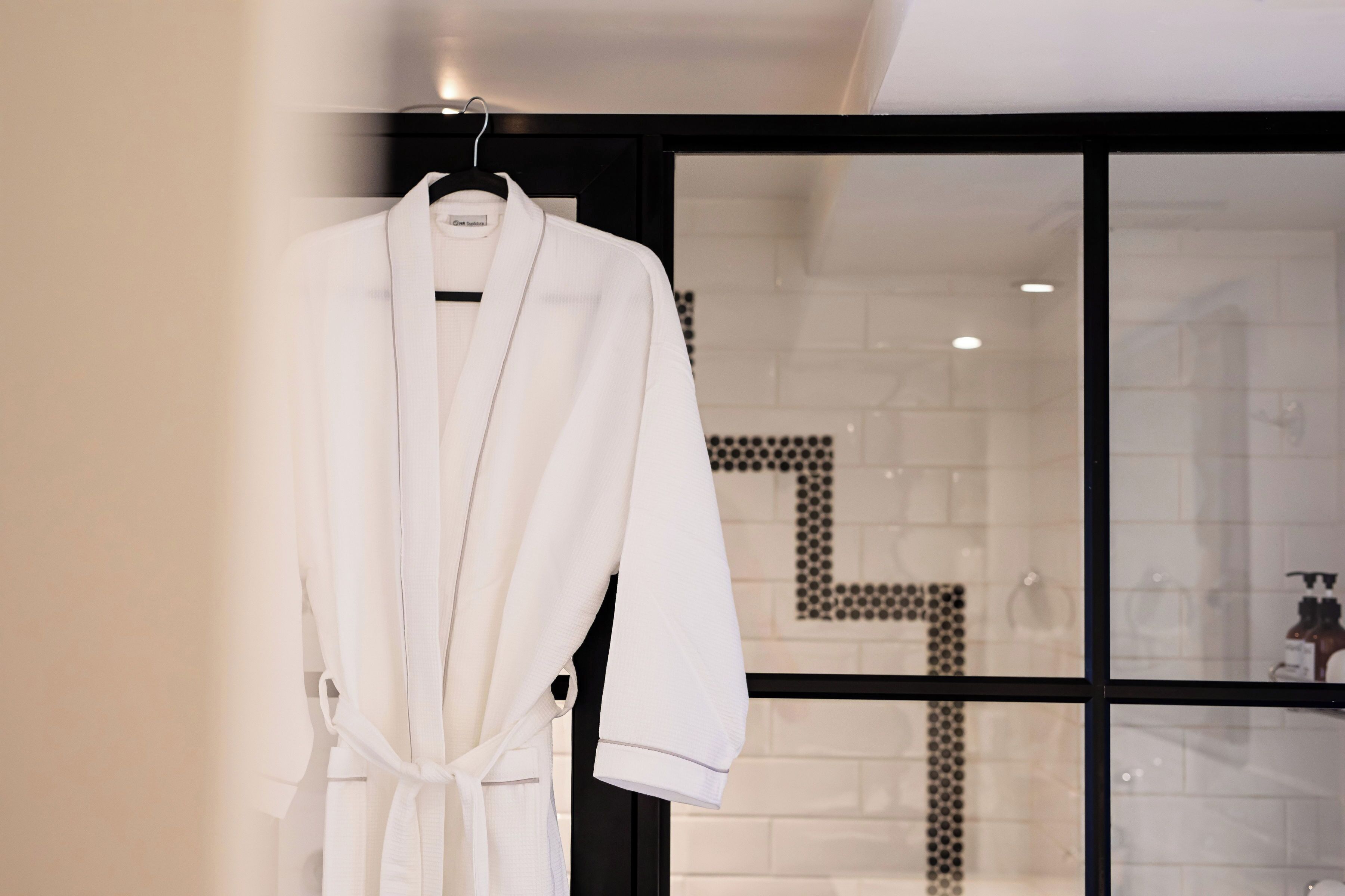 A white robe hanging on a hanger in a modern bathroom with a tiled shower area visible through a glass door.