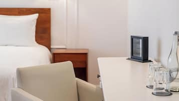 A hotel room with a bed, a chair, a small table with glasses and a picture frame, and a nightstand with a lamp.
