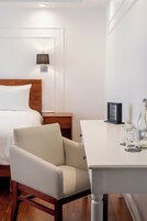 A hotel room with a bed, a chair, a small table with glasses and a picture frame, and a nightstand with a lamp.