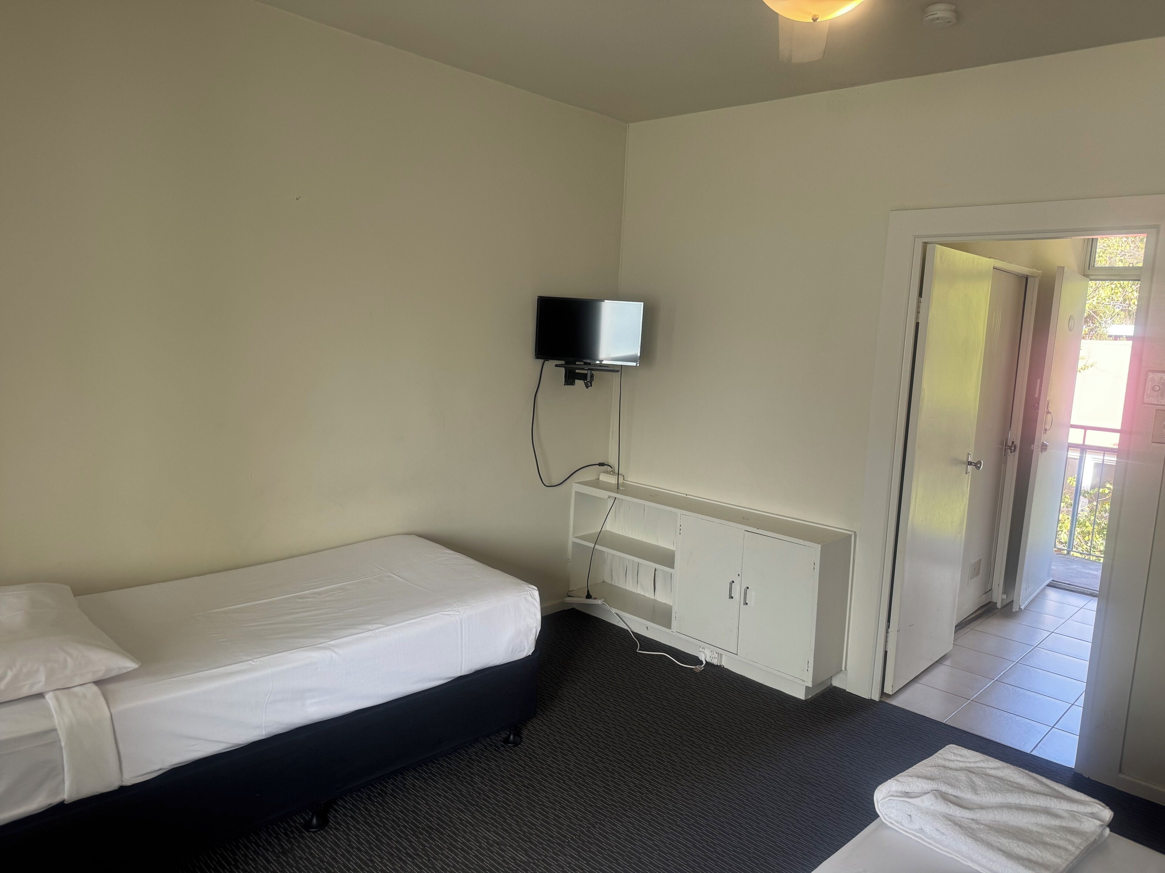 A single-bed room with a flat-screen TV mounted on the wall, a white wardrobe, and a door leading to an outdoor area.