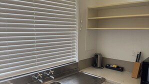 Una cocina con pileta de acero inoxidable, armarios blancos, una ventana con persianas y una encimera con hervidor y bloque de cuchillos.