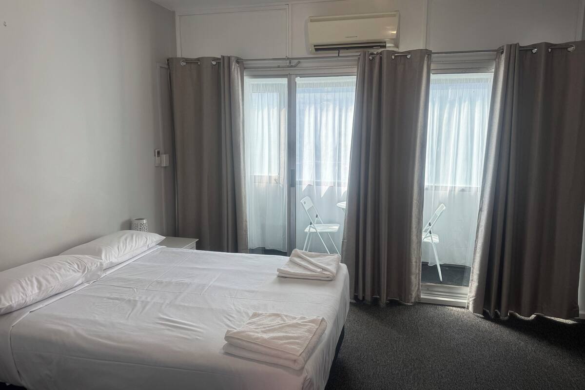 A hotel room with a bed, a window with curtains, a wall-mounted air conditioner, and a small table with a chair.
