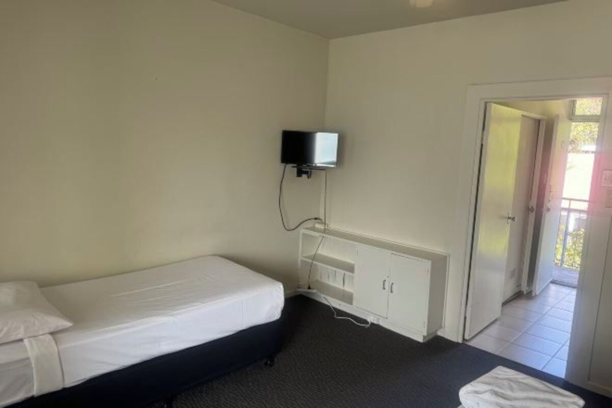 A single bed with a white mattress and a black headboard, a white cabinet below, and a television mounted on the wall.