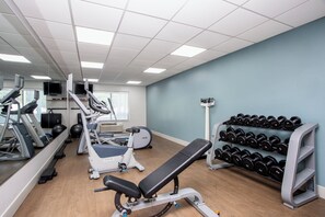 Academia com máquinas de cardio, um banco e uma estante com halteres.