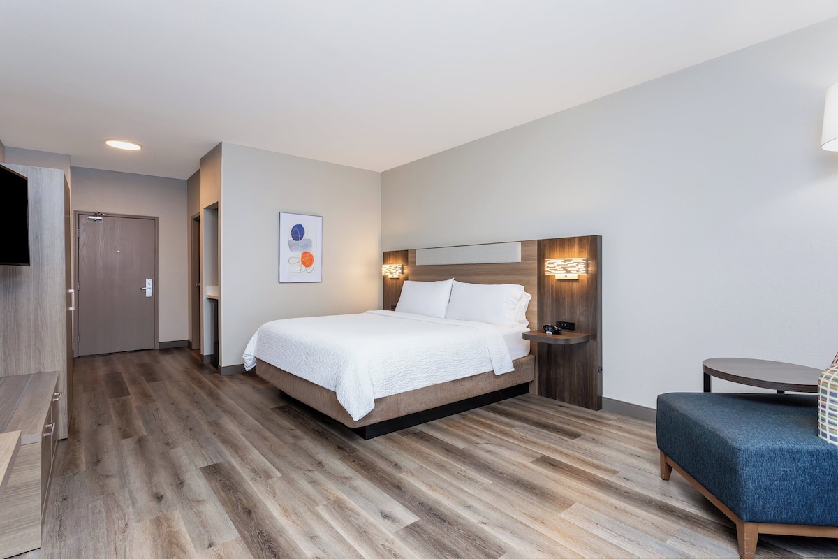 Quarto de hotel moderno com piso de madeira, cama com roupa de cama branca, mesa de cabeceira, mesa redonda pequena e sofá azul.