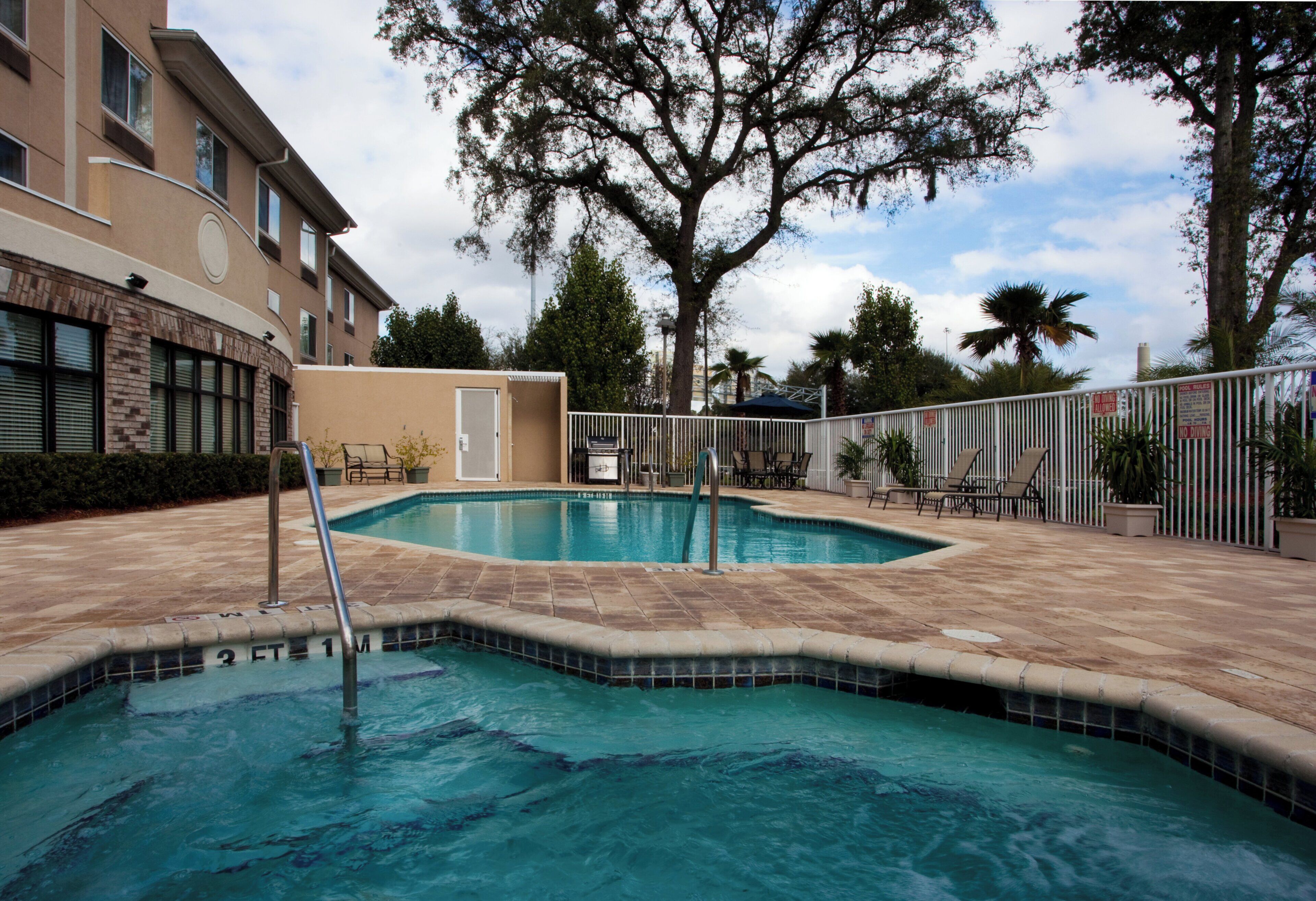 Una piscina con solárium de baldosas, rodeada por una valla y palmeras.