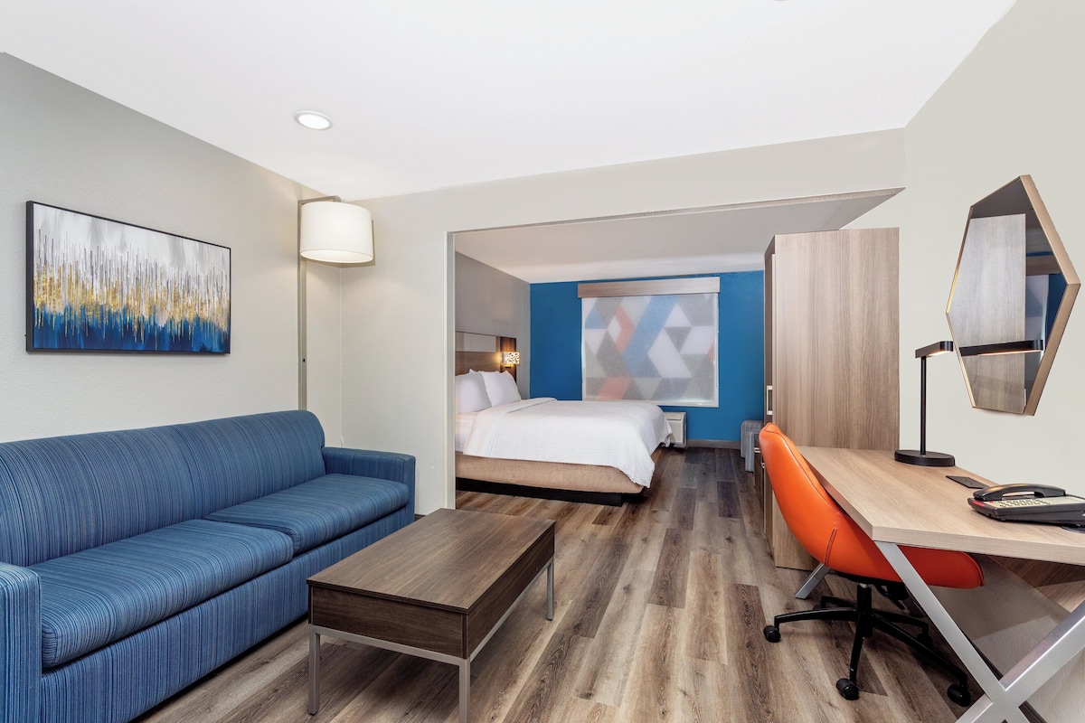 Quarto de hotel com um sofá azul, uma escrivaninha de madeira, uma cadeira de escritório laranja, uma cama com roupa de cama branca e um quadro abstrato grande na parede.