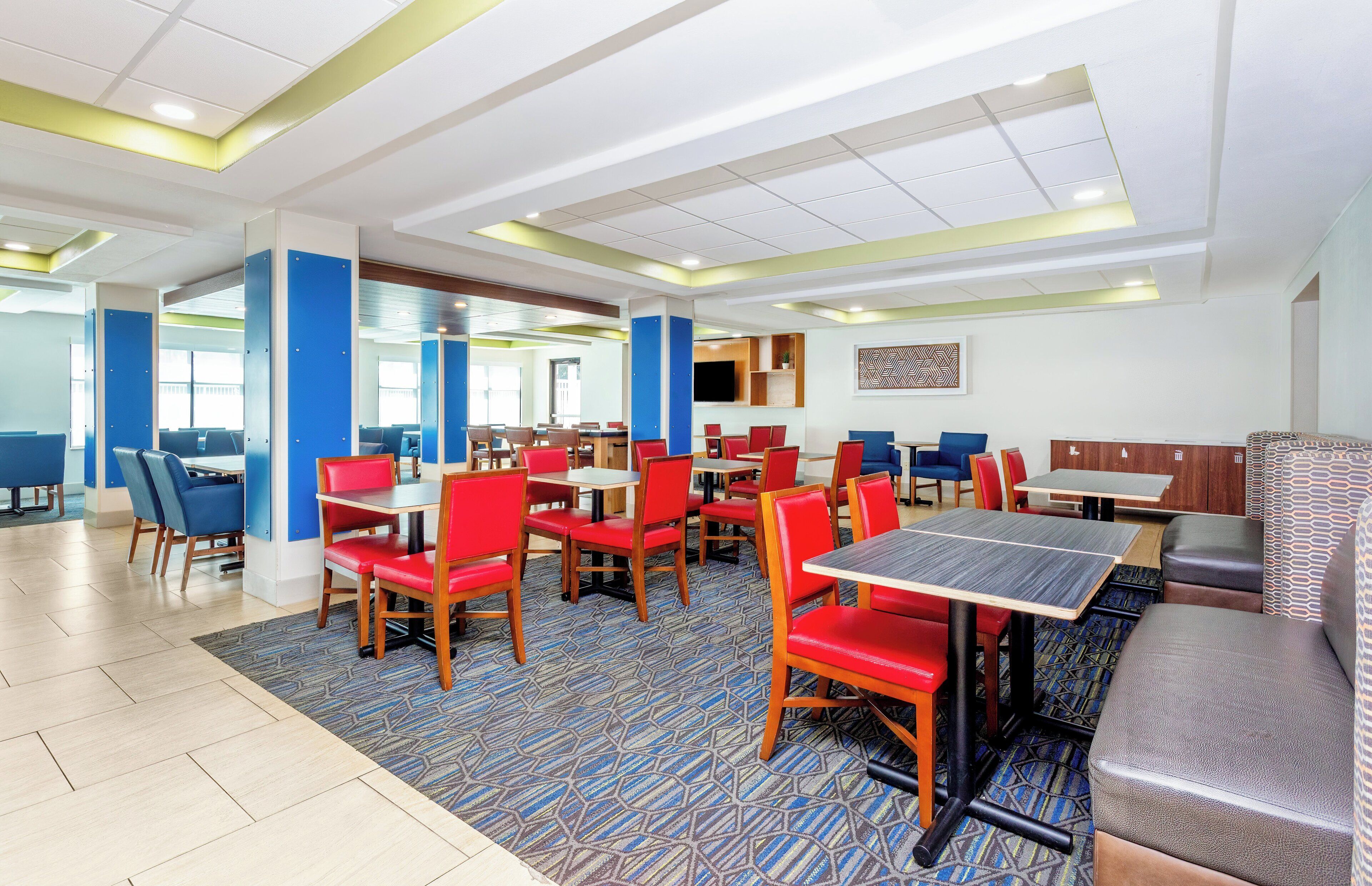 A hotel dining area with multiple tables and chairs, a patterned carpet, and a modern ceiling design.
