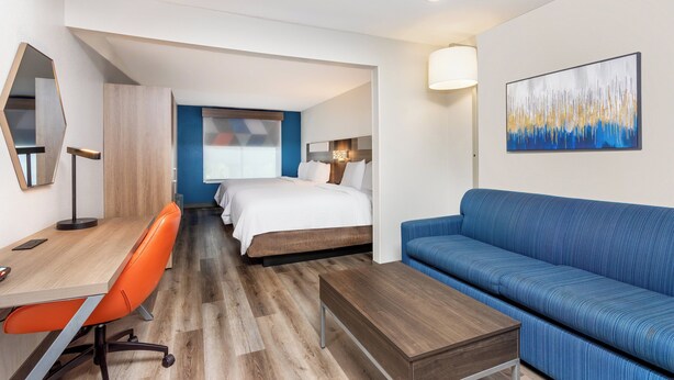 Une chambre d’hôtel avec un bureau en bois, une chaise orange, un canapé bleu et un lit avec une literie blanche.
