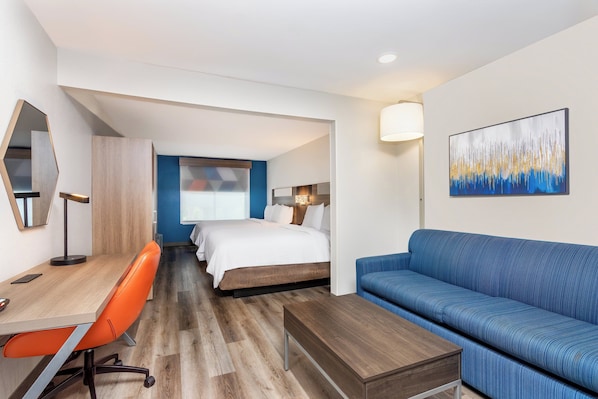 Quarto de hotel com escrivaninha de madeira, cadeira laranja, sofá azul e cama com roupa de cama branca.