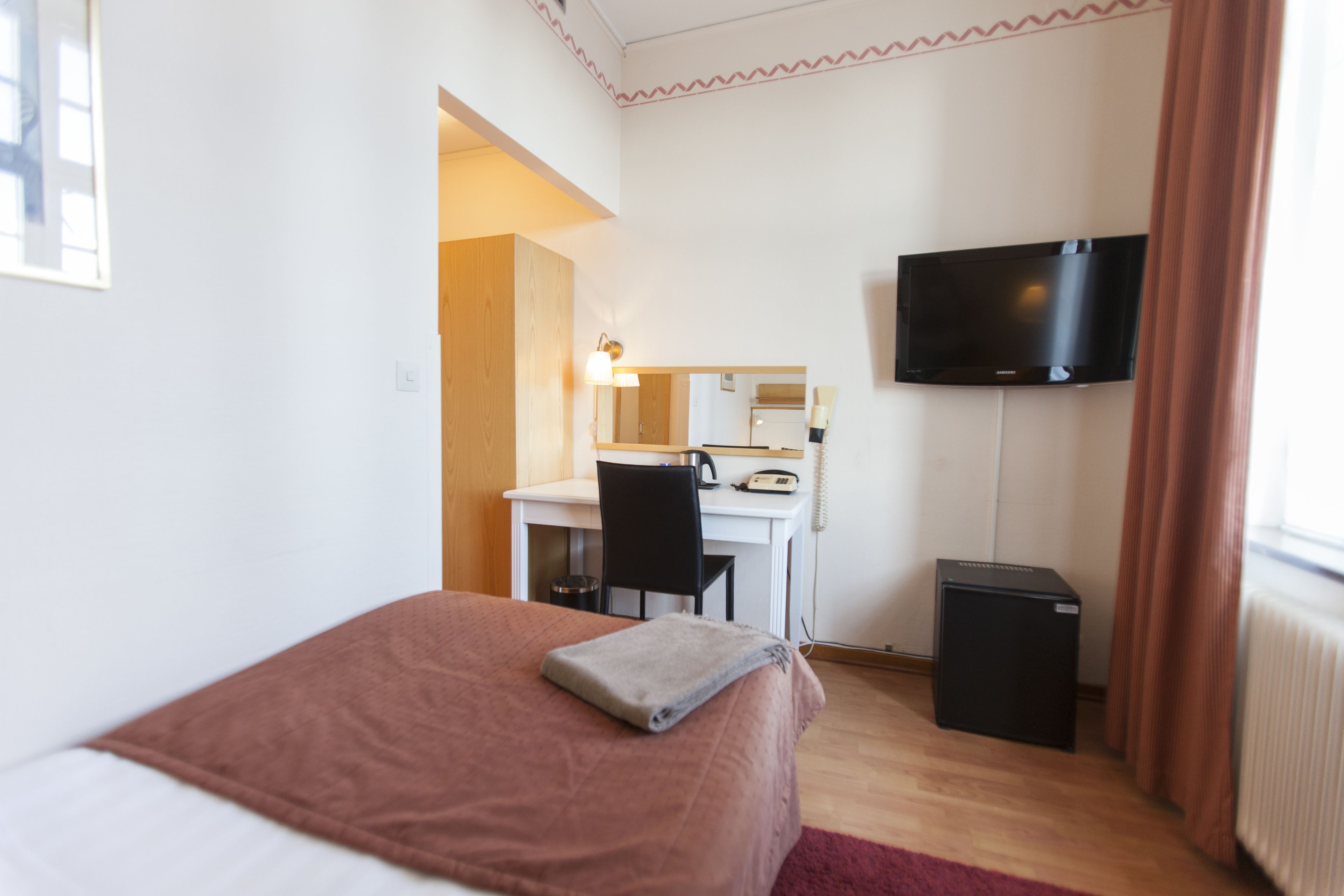 A hotel room with a bed, a desk, a chair, a television, and a small black cabinet.