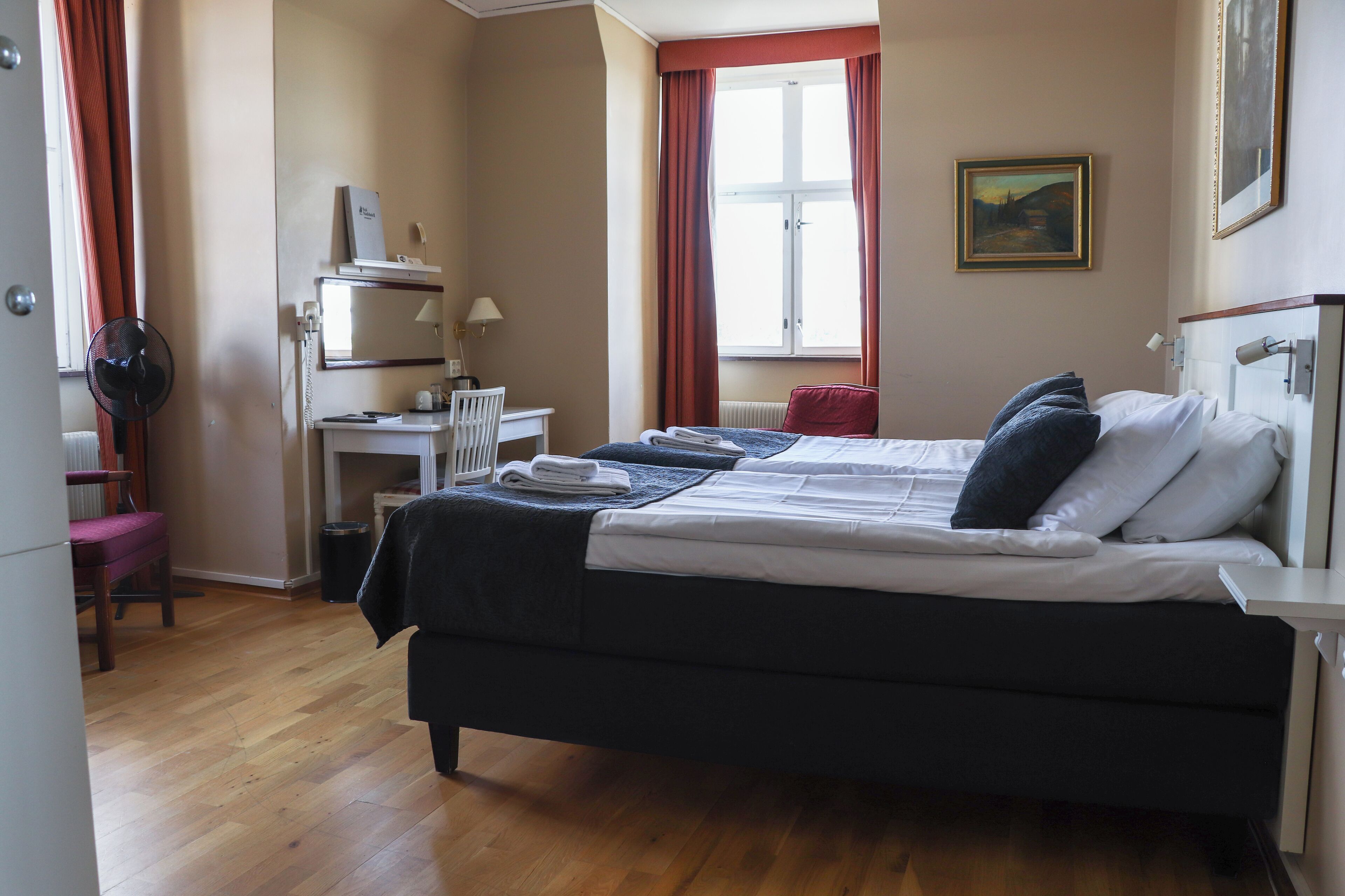 A hotel room with a bed, a desk, a chair, a radiator, and a window with curtains.