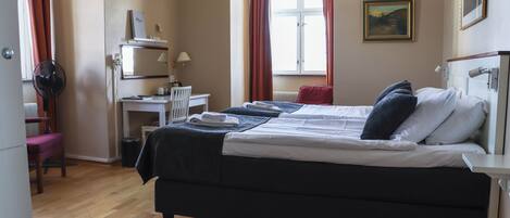 A hotel room with a bed, a desk, a chair, a radiator, and a window with curtains.