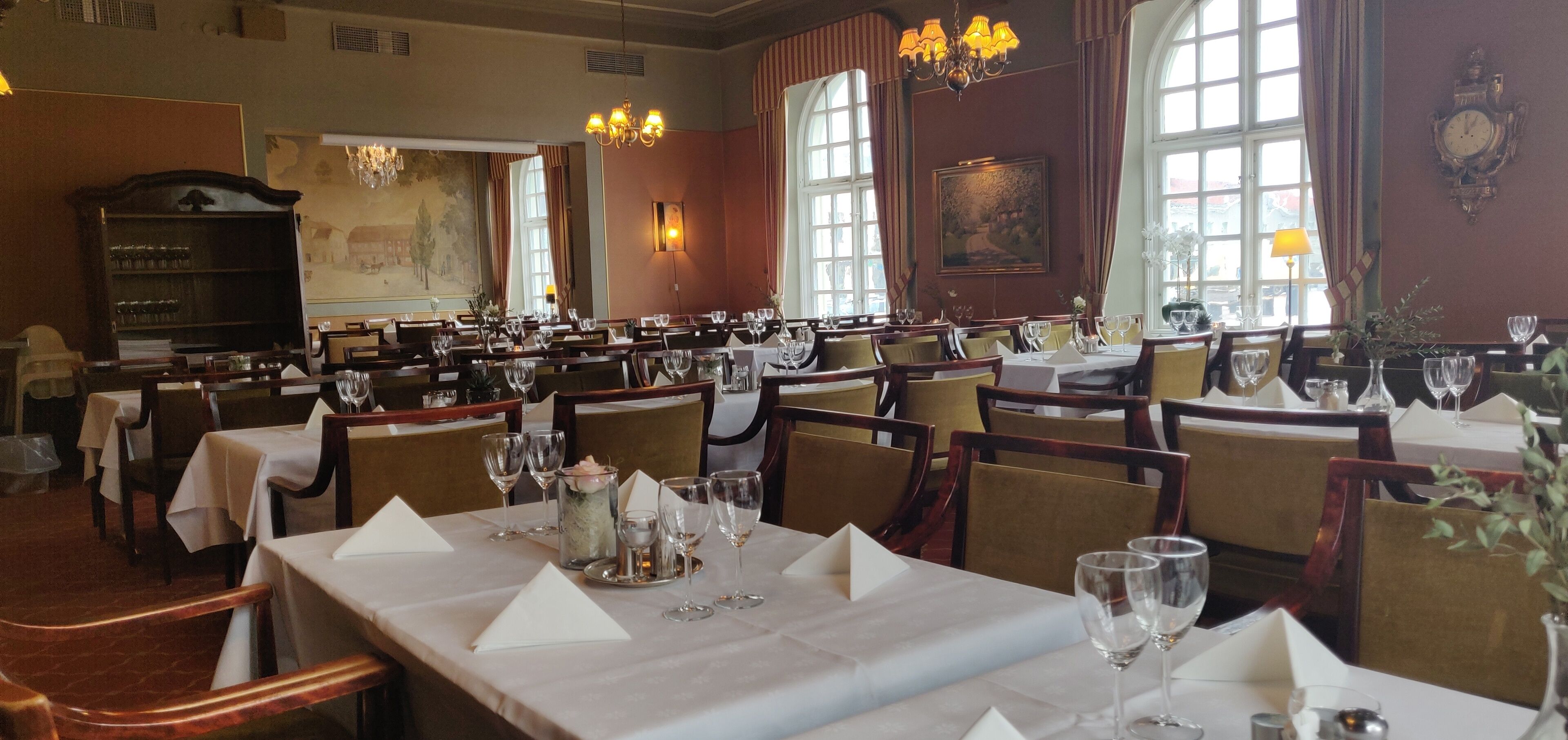 En restaurant med flere spiseborde, lysekroner og store vinduer.