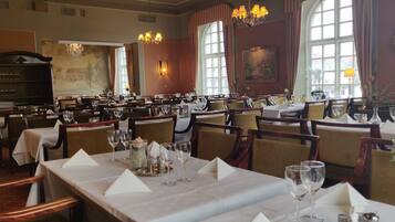 En restaurant med flere spiseborde, lysekroner og store vinduer.