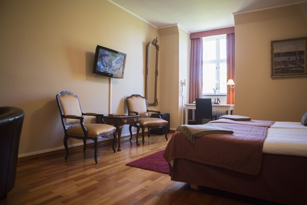 A hotel room with a wooden floor, two upholstered chairs, a bed with a red blanket, a TV mounted on the wall, and a window with curtains.